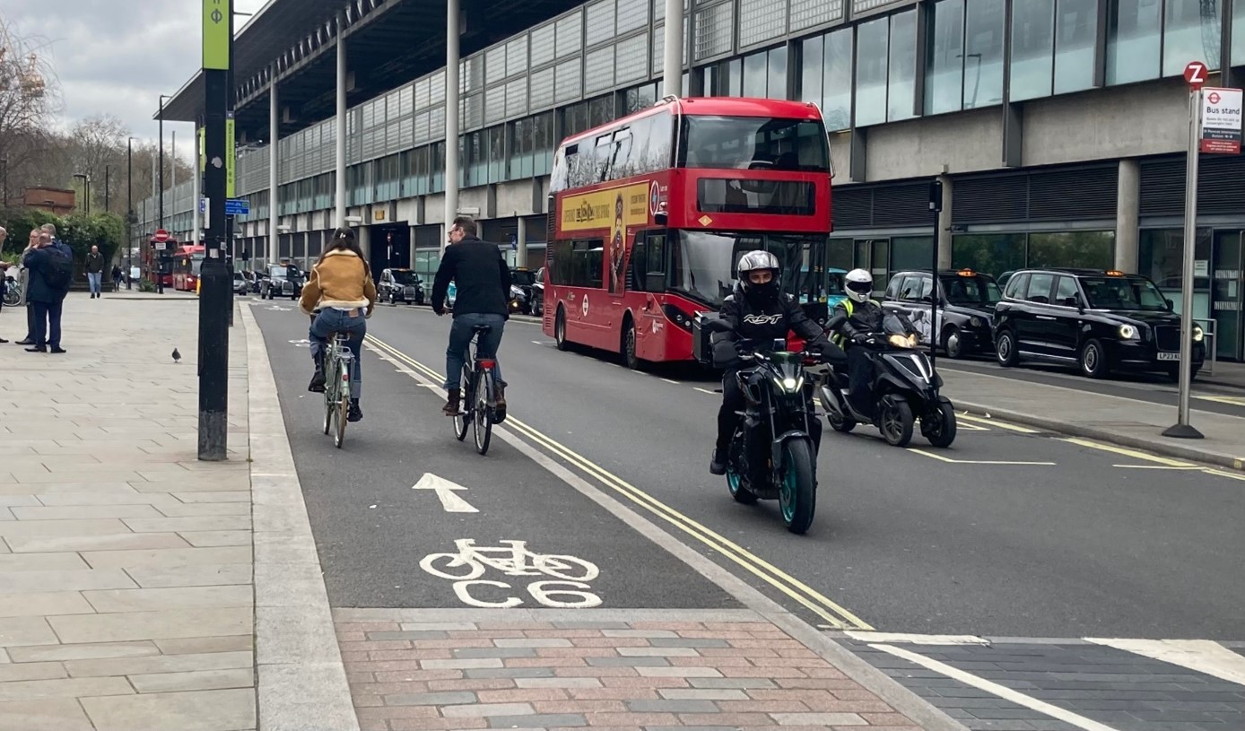 Image of road with pavement, cycle lane with cyclists and and one lane of motor traffic with motorbikes, cars and bus